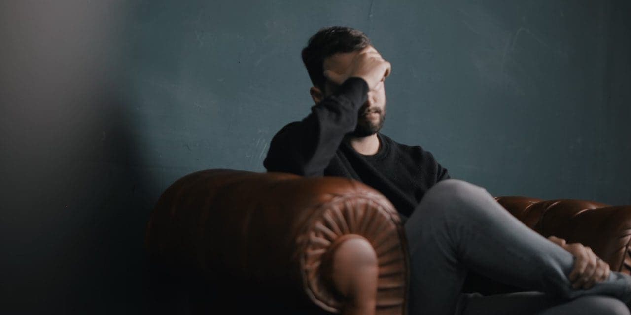 man sitting on couch, other leg on his lap while covering face with hand